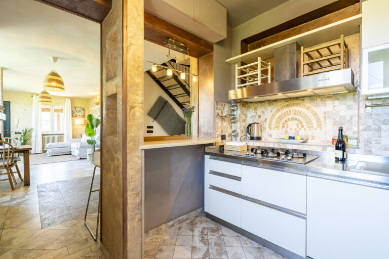 Cocina moderna con encimera de acero inoxidable, estufa a gas, decoración en azulejos y sala de estar con iluminación natural.