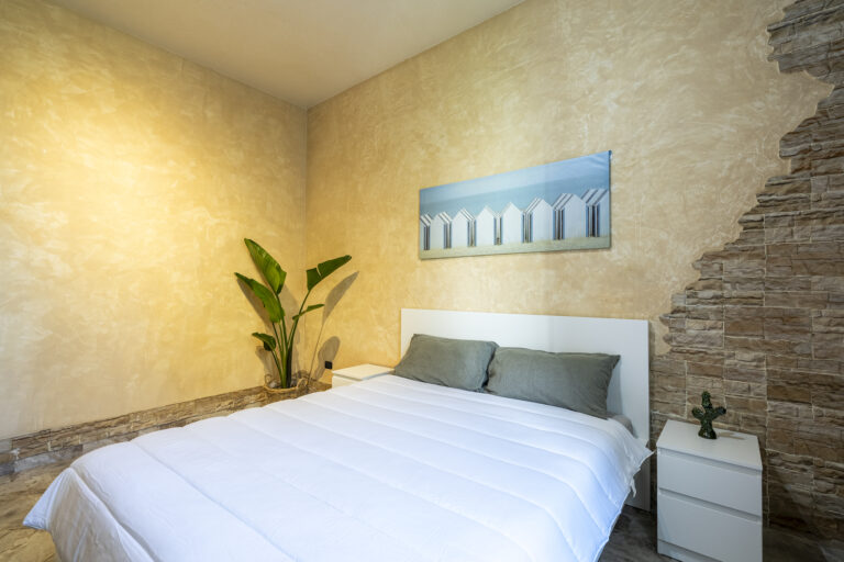 Dormitorio con cama blanca, dos cojines grises, pared en tonos beige, planta verde y foto de casetas azules. Mesa con cacto decorativo.