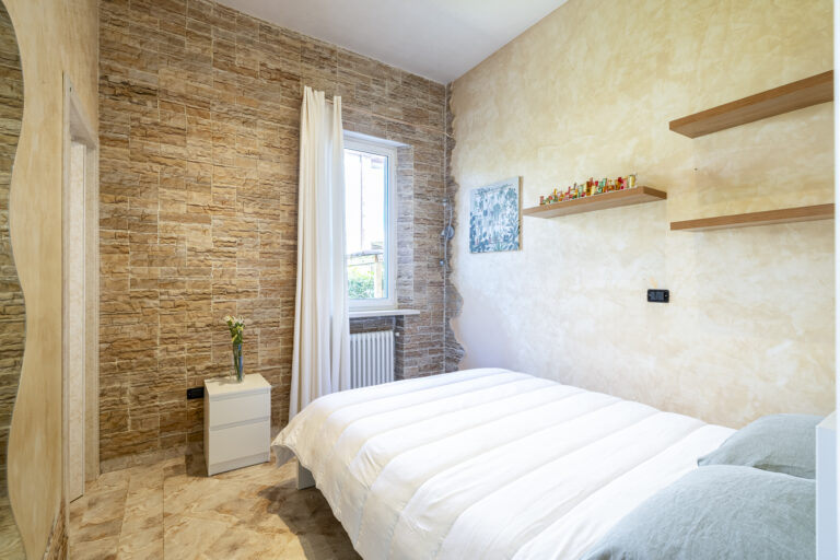 Dormitorio con paredes de piedra y beige, cama blanca, estante con decoraciones, ventana con cortina blanca y mesa de noche.