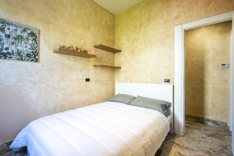 Habitación con paredes beige texturizadas, cama con cojines grises, estantes de madera y cuadro de plantas.