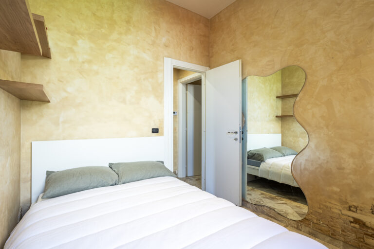 Habitación luminosa con paredes beige, cama con sábanas blancas, espejo ondulado y estanterías de madera en la pared.