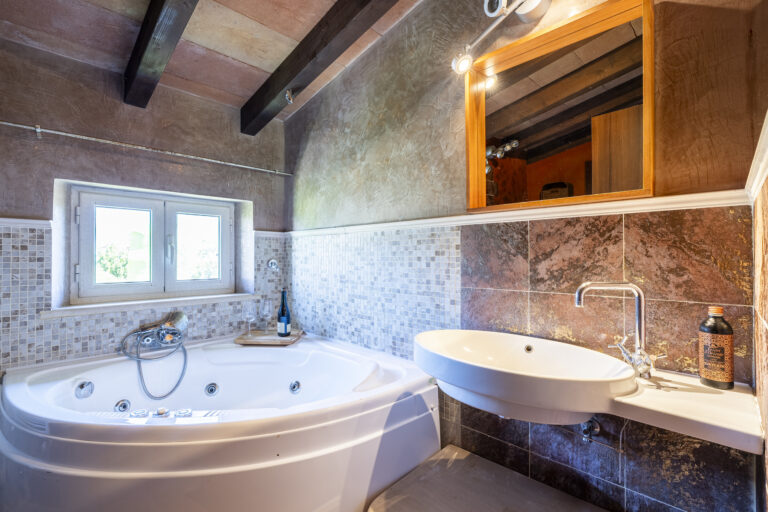 Baño con jacuzzi blanco, mosaico beige, lavabo moderno, espejo con luz. Botella y copas sobre borde. Ventana al exterior.