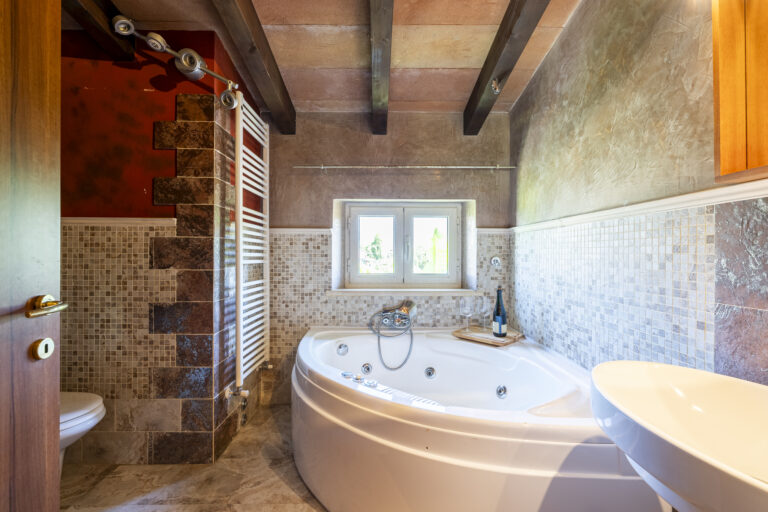 Baño elegante con jacuzzi, paredes de mosaico beige, ventana pequeña, techo con vigas y botella de vino con copas.