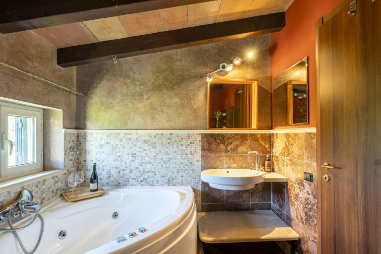 Baño con jacuzzi blanco, azulejos mosaico en beige, lavabo blanco, espejo, pared de tonos tierra y techo con vigas de madera.