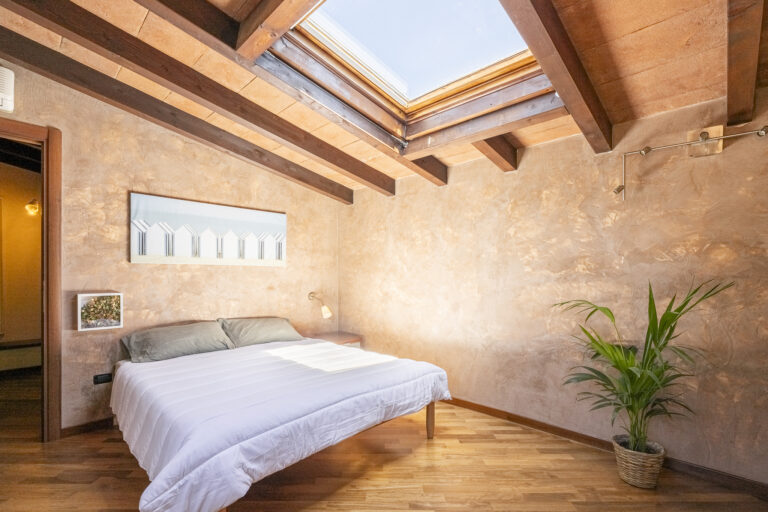 Dormitorio con techo de vigas y tragaluz, cama doble, pared texturizada beige, planta en maceta y arte minimalista.