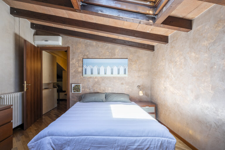 Habitación con cama doble, vigas de madera, pared beige texturizada, arte moderno, ventana en el techo y lámpara de pared.