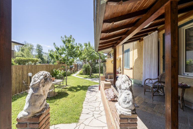 Casa con porche de madera, estatuas de leones, camino de piedra y jardín verde con árboles. Sillas en el porche.