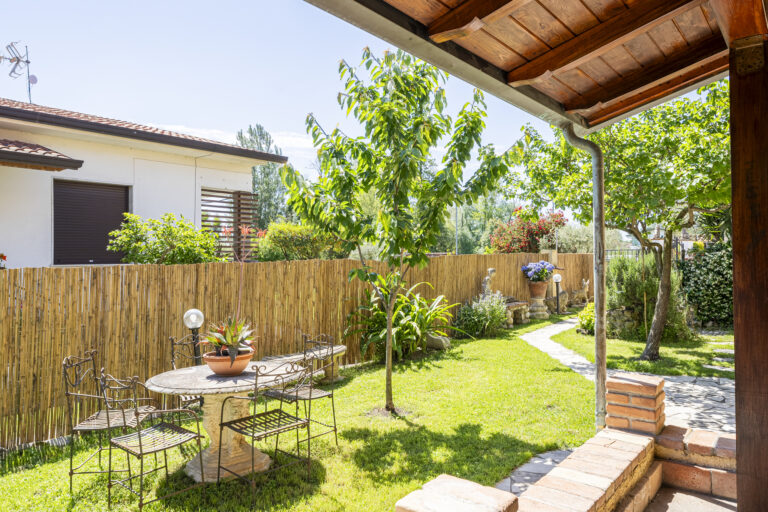 Jardín con césped verde, mesa redonda de piedra, sillas de metal, plantas, árboles y cerca de bambú bajo techo de madera.