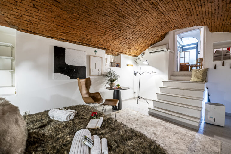 Espacio acogedor con techo de ladrillo, silla moderna, mesa redonda con planta, escalera blanca y alfombra peluda.