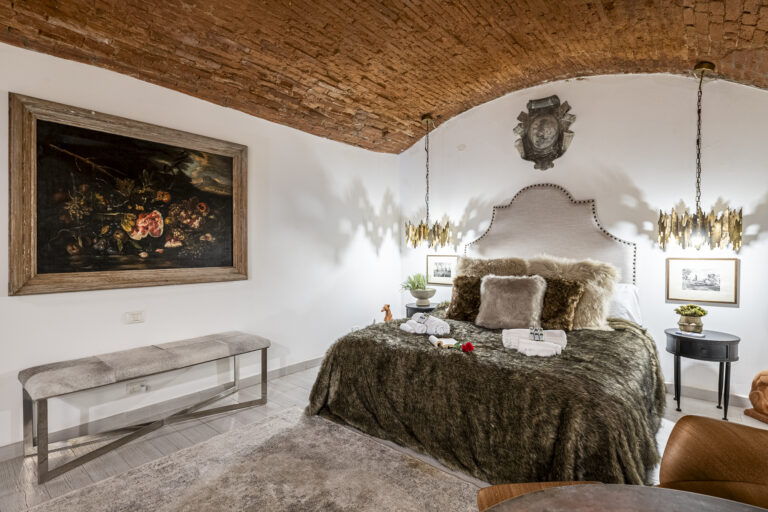 Dormitorio elegante con techo abovedado de ladrillo, cama con mantas de piel, lámparas doradas, y arte clásico en paredes blancas.