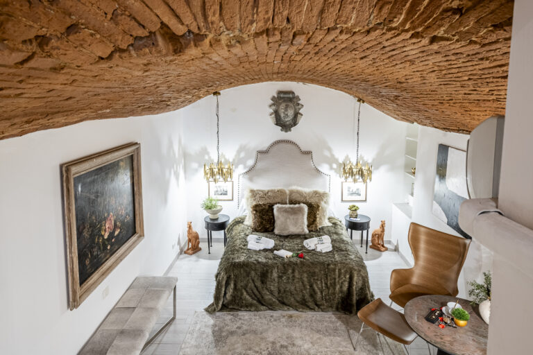 Habitación con techo abovedado de ladrillo, cama con cobertor de piel, lámparas doradas, sillas modernas y decoración clásica.