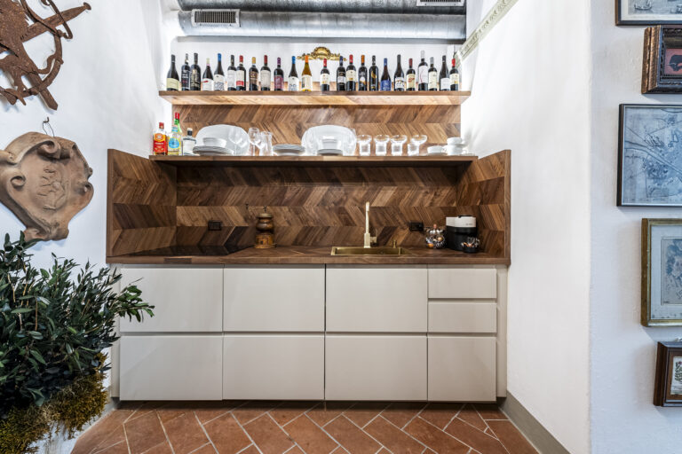 Cocina con estantes de madera, botellas de vino, platos, copas, fregadero dorado. Decoración de pared y planta en esquina.