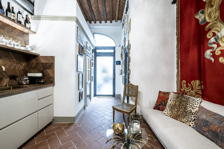 Entrada con piso de baldosas, cocina de madera, sofá con cojines estampados, silla de mimbre, y puerta de vidrio esmerilado.