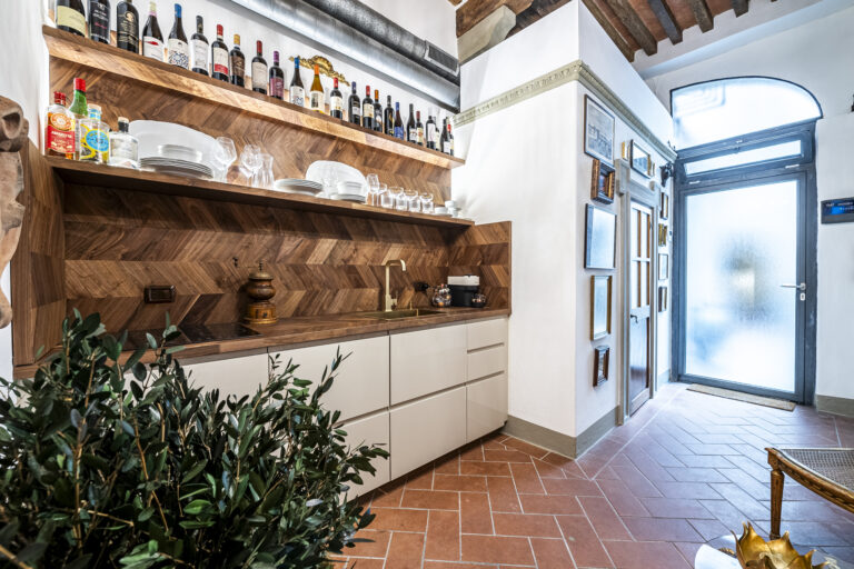 Cocina moderna con estantes de madera, botellas de vino, platos, vasos y una planta. Puerta de cristal en el fondo.