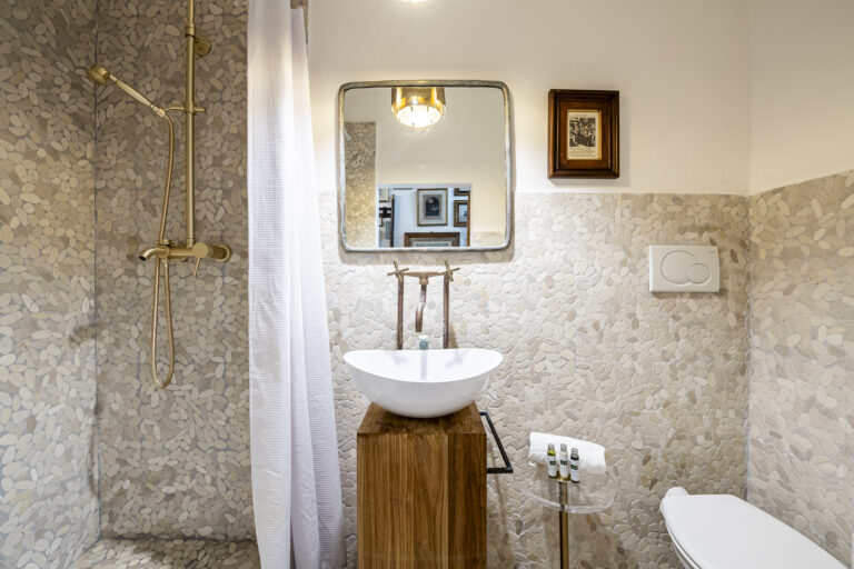 Baño con ducha de mosaico de piedra, lavamanos sobre mueble de madera, espejo cuadrado y detalles en tonos neutros.