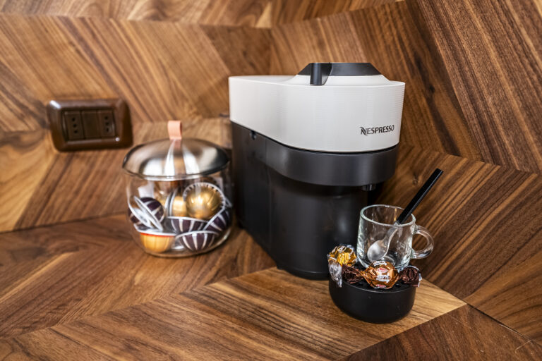 Máquina de café Nespresso en mostrador de madera, cápsulas en frasco, taza de vidrio con cuchara y chocolates envueltos al lado.