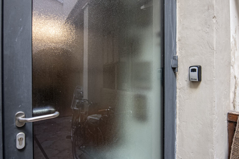 Puerta de vidrio opaco con marco gris y manija metálica. Al lado, una caja segura montada en la pared y una bicicleta reflejada.