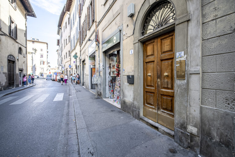 Calle urbana con edificios antiguos, una tienda de souvenirs, puerta de madera con herrajes dorados y personas caminando.