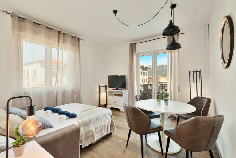 Sala luminosa con cama, mesa redonda blanca y sillas marrones. Ventanas grandes con cortinas beige. TV y decoración moderna.