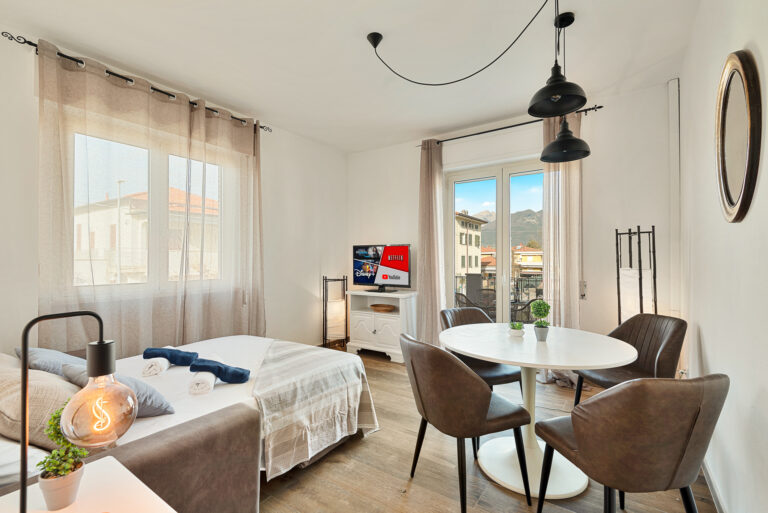 Sala luminosa con cama, TV plana, mesa redonda y cuatro sillas. Ventanas grandes con cortinas, lámpara colgante y decoración moderna.