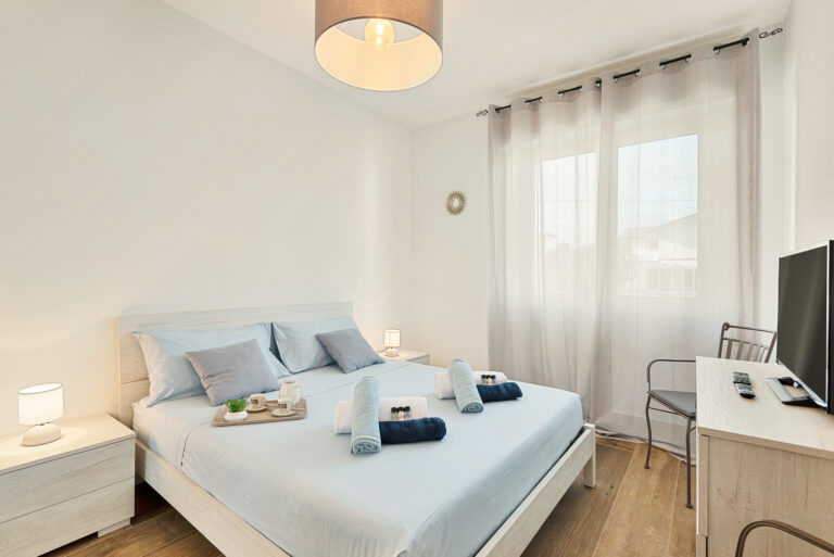 Habitación luminosa con cama doble, sábanas azules y lámparas en las mesitas. Ventana con cortinas translúcidas. Televisor sobre mesa.