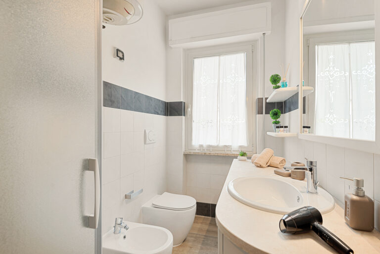 Baño moderno con lavabo blanco, inodoro, bidé, espejo grande, y ducha. Decorado con plantas, toallas y cortinas blancas.