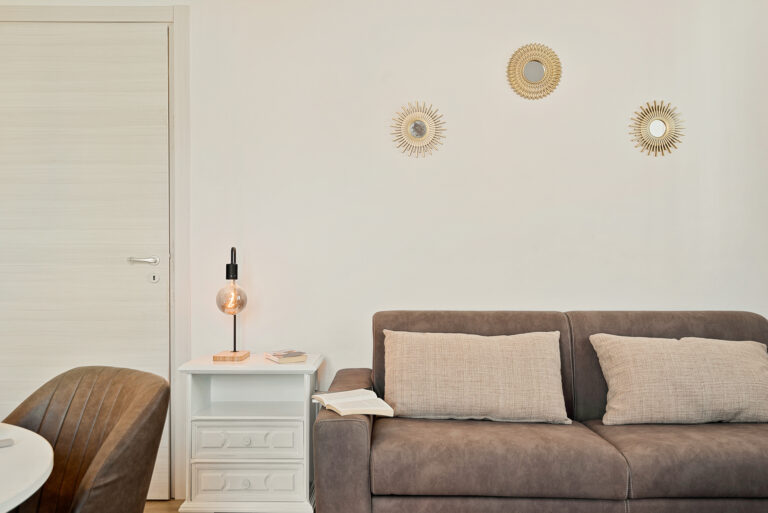 Sofá marrón con cojines beige, lámpara de mesa encendida, pared blanca con tres espejos decorativos. Puerta a la izquierda.