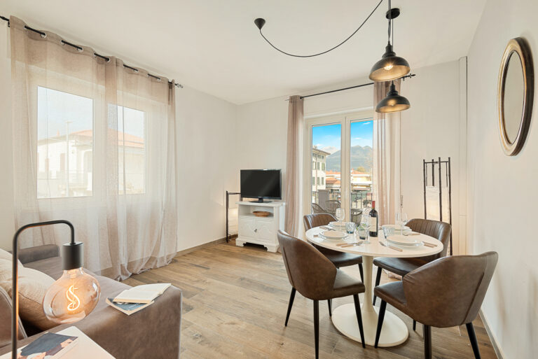 Sala moderna con suelo de madera, mesa redonda con sillas, lámparas colgantes, ventanales con cortinas y televisor en mueble blanco.