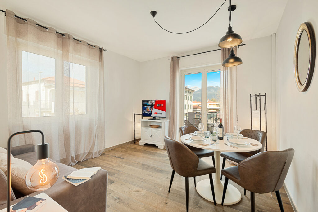 Sala moderna con piso de madera, mesa redonda para cuatro personas, sofá gris, televisor y ventanas grandes con cortinas claras.