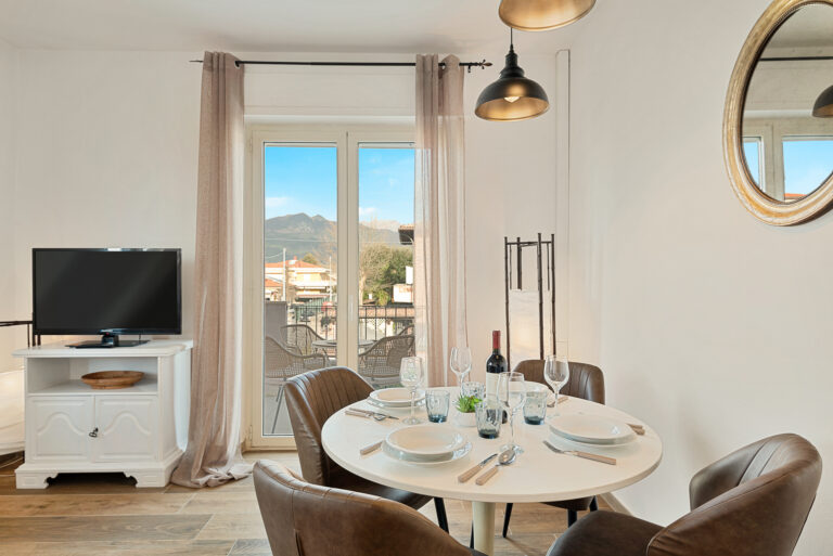 Sala de estar con mesa redonda, sillas marrones, TV sobre mueble blanco, ventana a balcón con vistas a montañas.