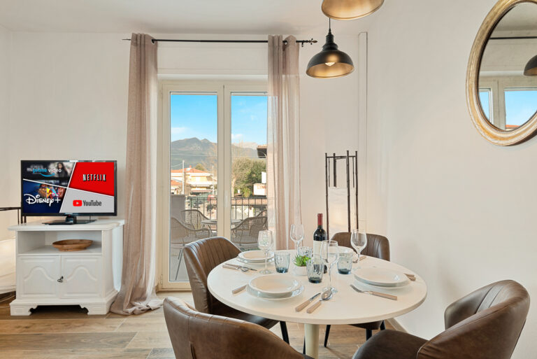 Sala de estar con mesa puesta, sillas marrones, ventana panorámica, televisor y luces colgantes. Vista de montañas al fondo.