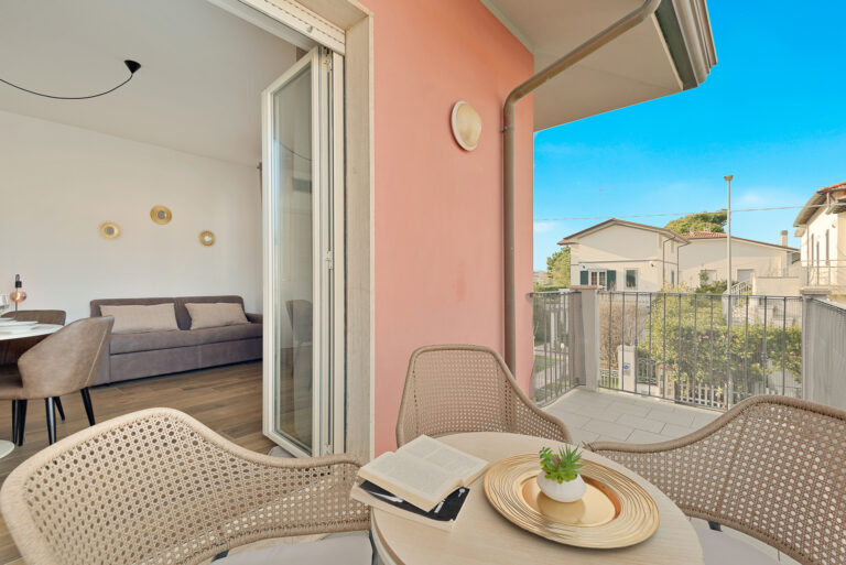 Balcón con sillas de mimbre y mesa, libro abierto y planta decorativa. Interior moderno con sofá gris y cuadros dorados. Vista a casas vecinas.