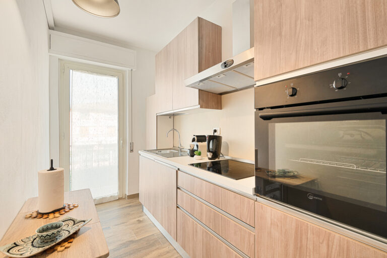 Cocina moderna con muebles de madera clara, horno, vitrocerámica, cafetera, fregadero y ventana grande. Decoración minimalista.