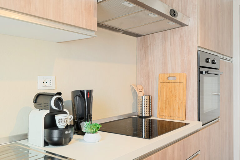 Cocina moderna con electrodomésticos negros, cafetera, tetera y placa de inducción en encimera de madera clara.