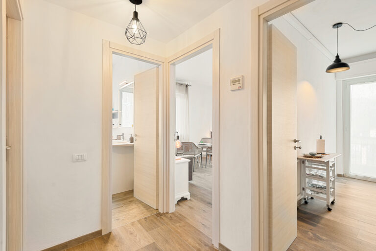 Pasillo con suelo de madera, tres puertas abiertas; una al baño, otra a la sala con muebles y ventana, y otra a la cocina con carrito blanco.