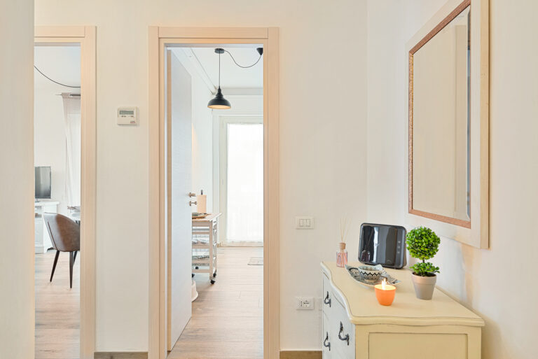 Pasillo de casa con dos puertas abiertas, mueble de cajones, espejo, planta decorativa, vela encendida y suelo de madera clara.