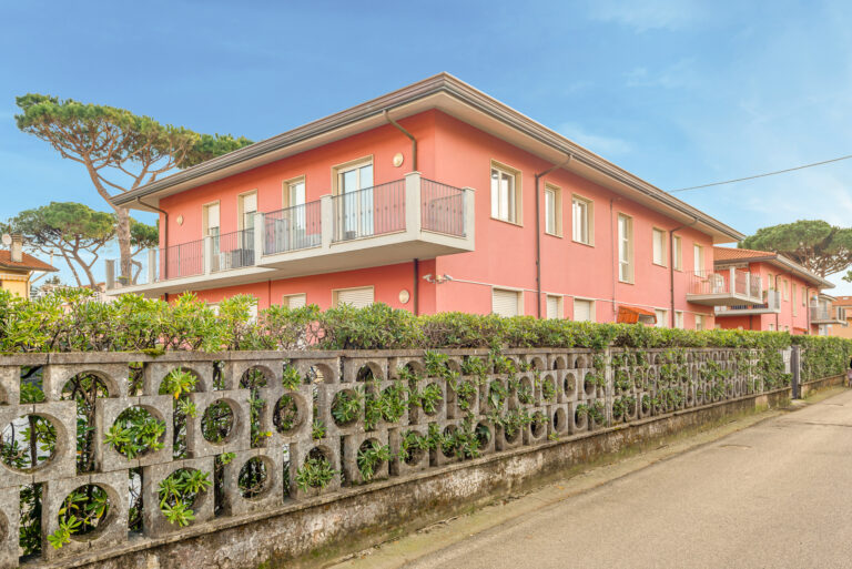 Edificio de dos pisos con fachada rosada, balcones y ventanas. Cercado de concreto con arbustos verdes y árboles altos al fondo.