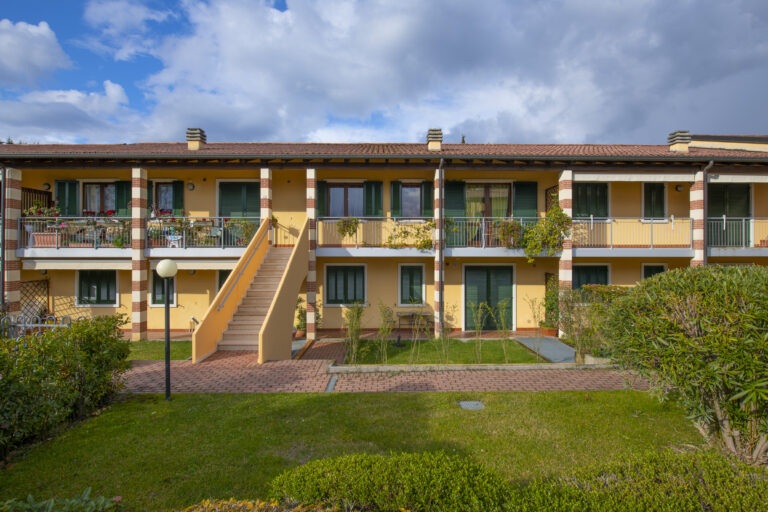 Edificio de dos pisos con balcones y ventanas verdes. Jardín delantero, escalera central y cielo nublado.
