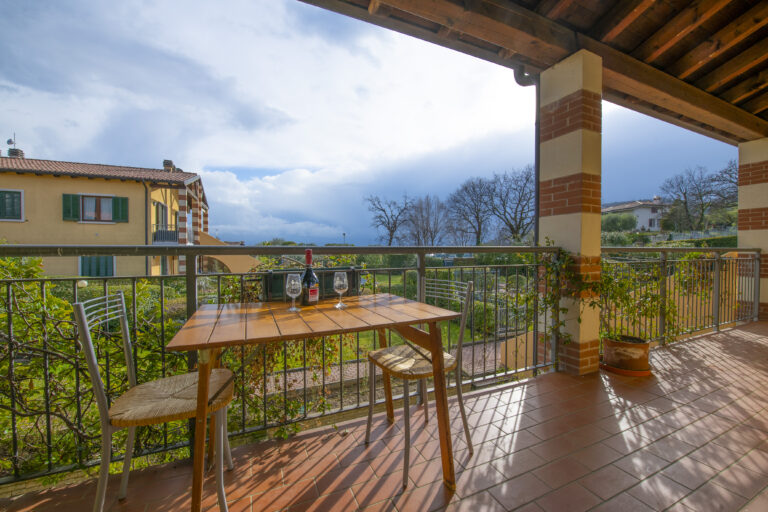 Terraza con mesa de madera, dos sillas, botella de vino y copas. Vista de jardín verde, árboles y cielo nublado.