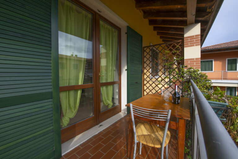 Balcón acogedor con muebles de madera, sillas de metal, cortinas verdes y vistas a edificios vecinos.