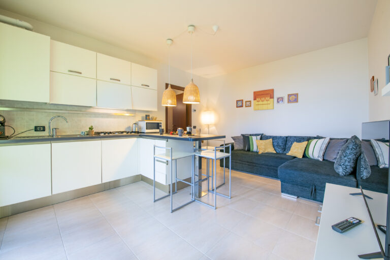 Cocina moderna en blanco con encimera oscura, lámparas colgantes, sofá azul con cojines coloridos y decoración de pared.