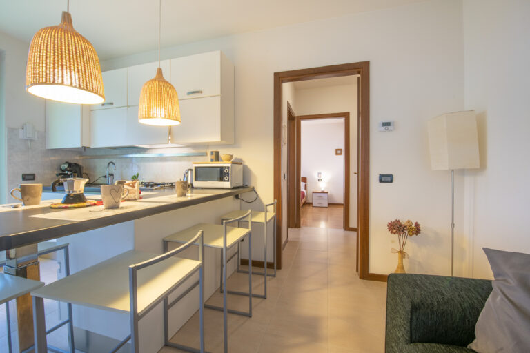 Cocina moderna con lámparas colgantes tejidas, encimera con tazas, microondas, y taburetes. Al fondo, entrada a dormitorio.