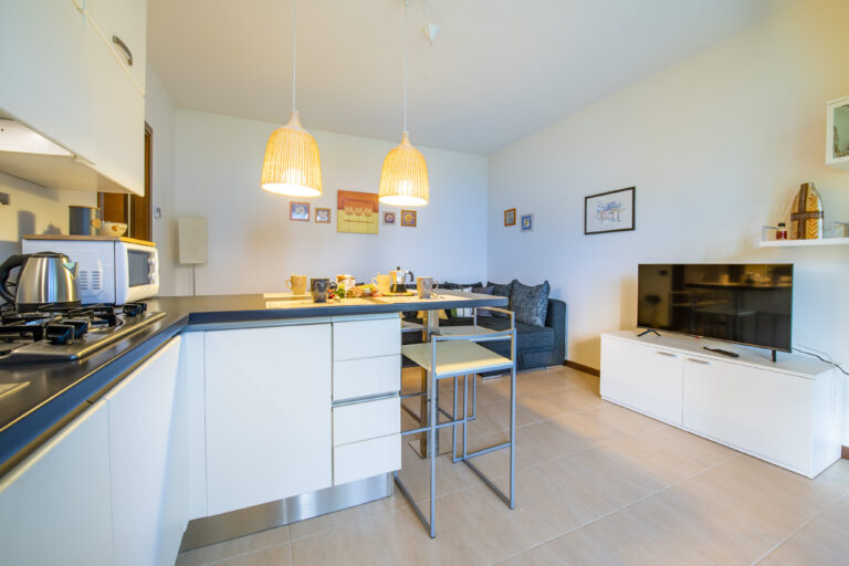 Cocina moderna con encimeras negras, taburetes blancos, lámparas colgantes y sala de estar con sofá y televisor en mueble blanco.