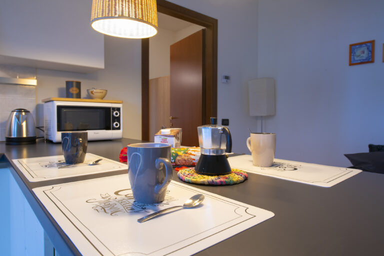 Mesa de cocina con tazas, cafetera moka, microondas, tetera y decoración acogedora. Iluminación cálida colgante.