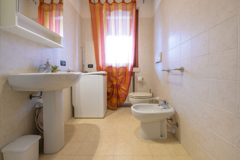 Baño con lavabo, lavadora, inodoro y bidet. Cortina naranja en la ventana, azulejos beige y suelo de cerámica.