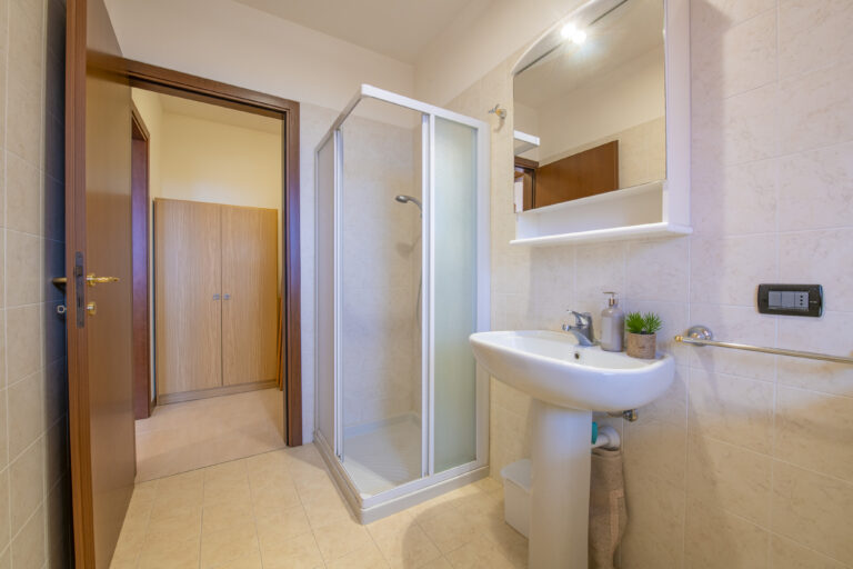 Baño con ducha de mampara, lavabo con espejo, toallero y luz tenue. Puerta abierta muestra armario en pasillo.