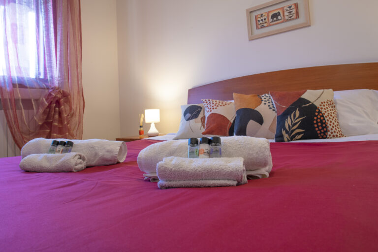 Habitación con cama doble, sábanas rojas, cojines coloridos, toallas blancas con botellas de aseo y una lámpara de mesa.