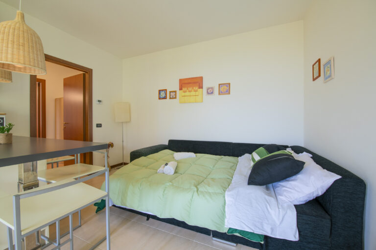 Habitación moderna con sofá cama oscuro, edredón verde, lámpara de pie, cuadros pequeños en la pared y mesa con sillas.