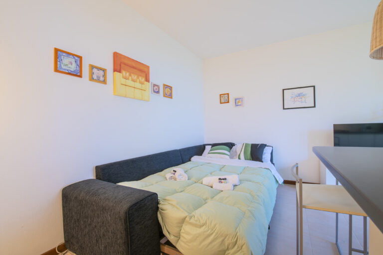 Sofá cama gris oscuro con edredón verde, cojines a rayas. Pared blanca decorada con cuadros. Mesa alta con sillas y televisor.