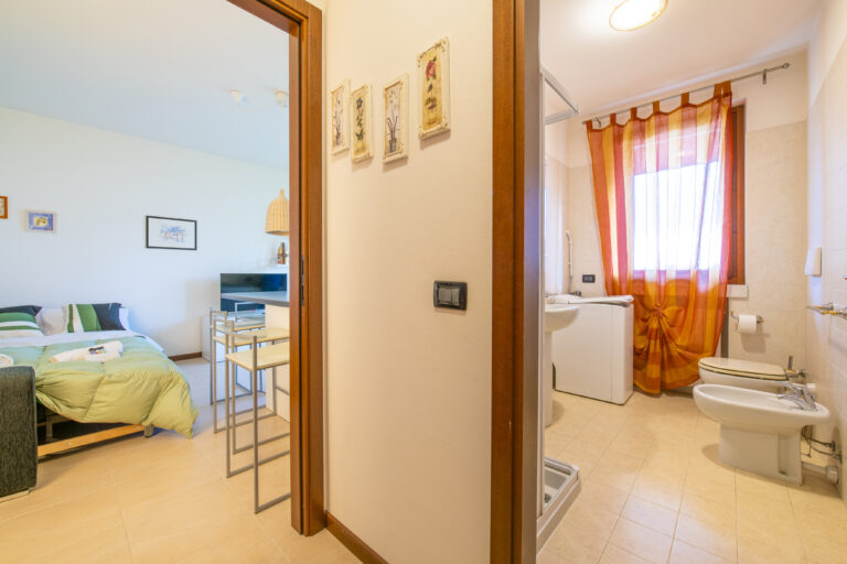 Habitación con cama matrimonial, edredón verde y paredes con cuadros. Baño con bidé, cortinas naranjas y lavamanos.